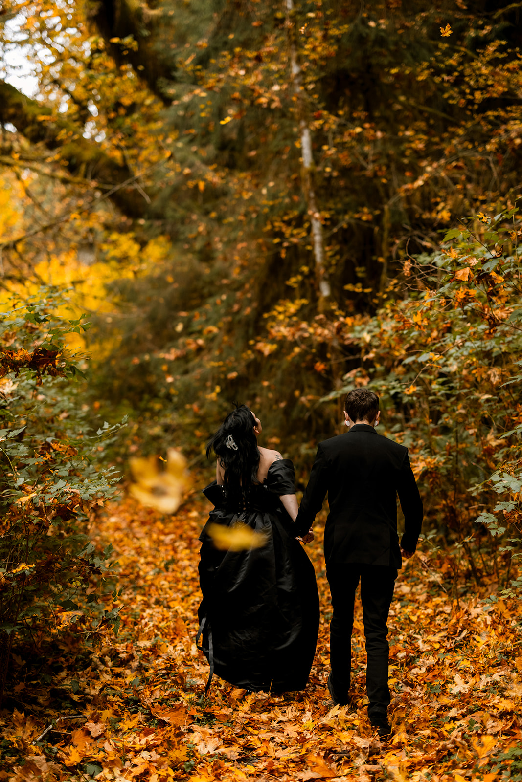 A couple in black elopement outfits walk hand in hand away from the camera, as orange leaves fall and cover the ground.