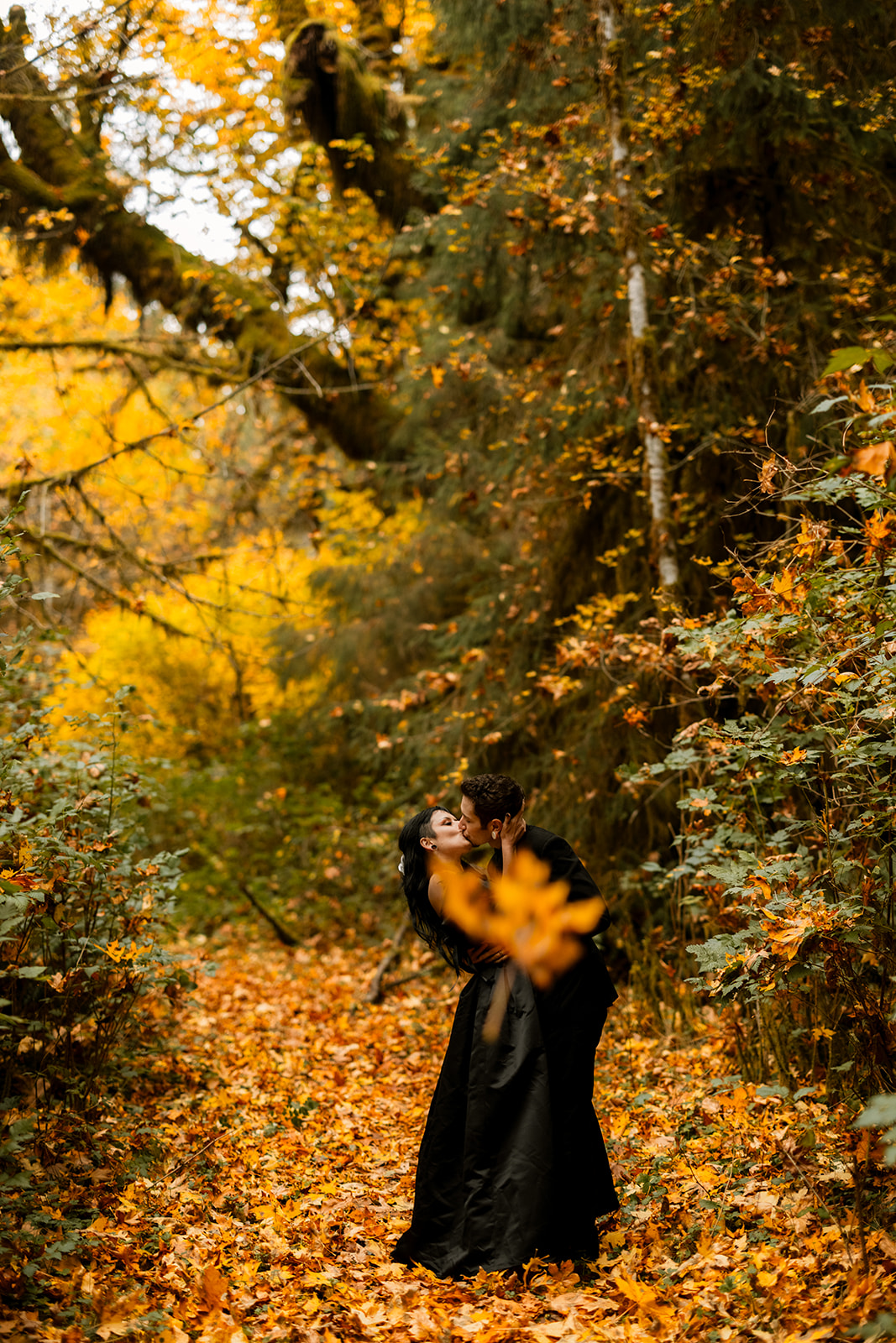 A couple in black elopement outfits share a dip kiss, as orange leaves fall and cover the ground.