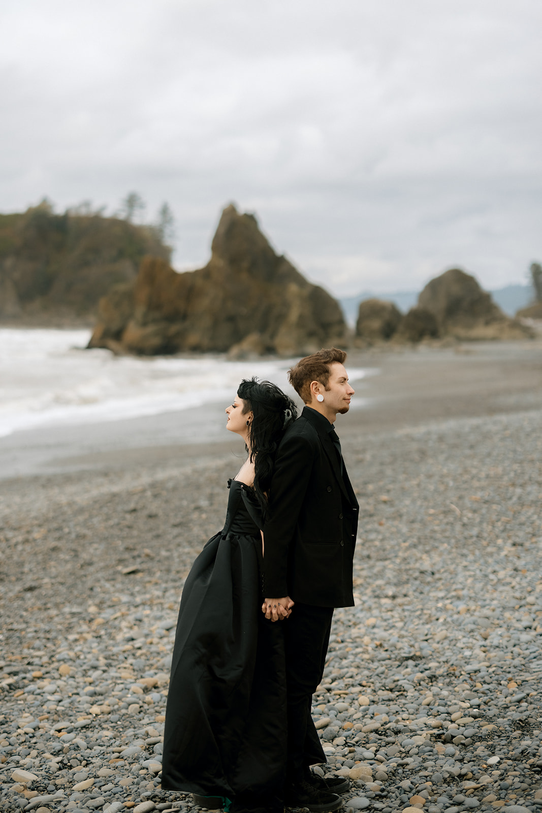 A couple in black elopement outfits stand back to back on a Washington beach during their first look at their wedding.