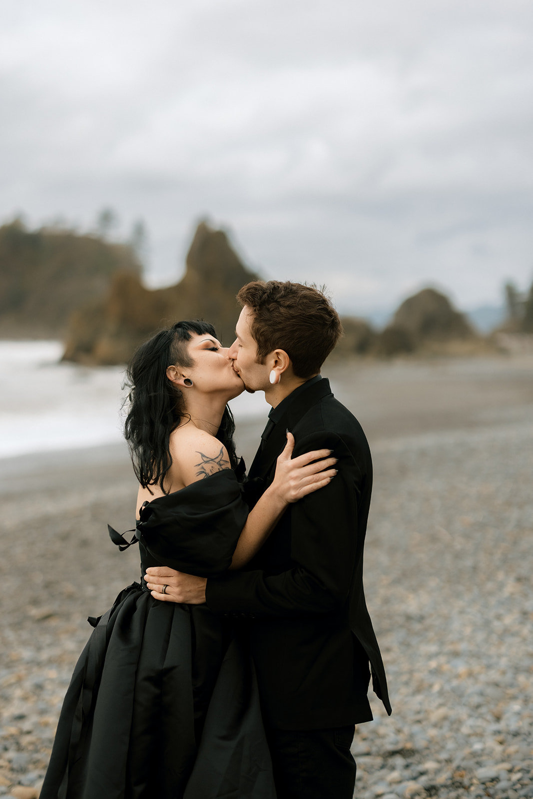 A couple in black wedding outfits kisses on a washington beach on a cloudy day.