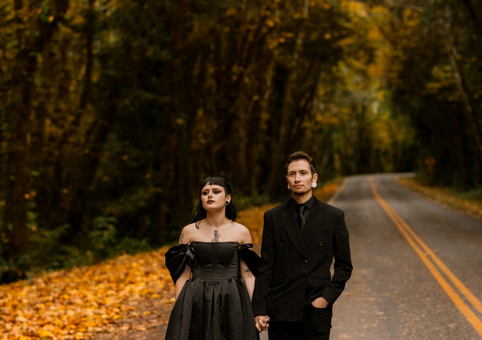 Two people in black wedding outfits walk hand in hand on a blacktop road, with fall leaves falling to the ground.