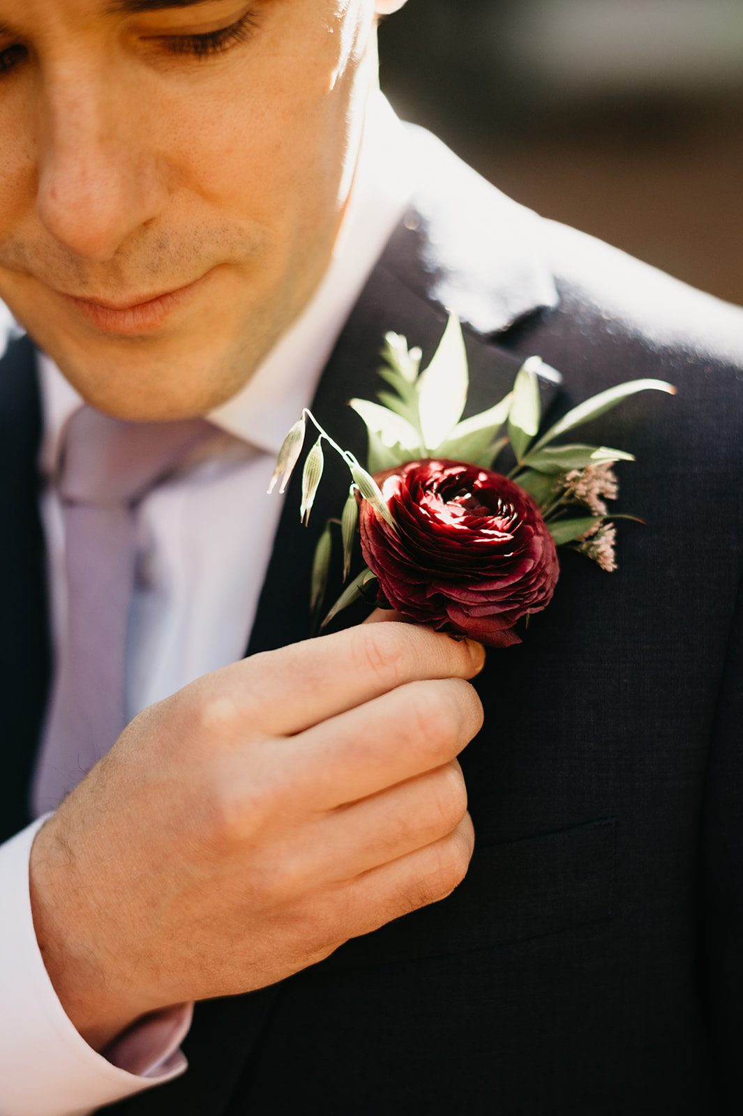 The groom in a dark suit puts his boutonniere on his suit coat. 