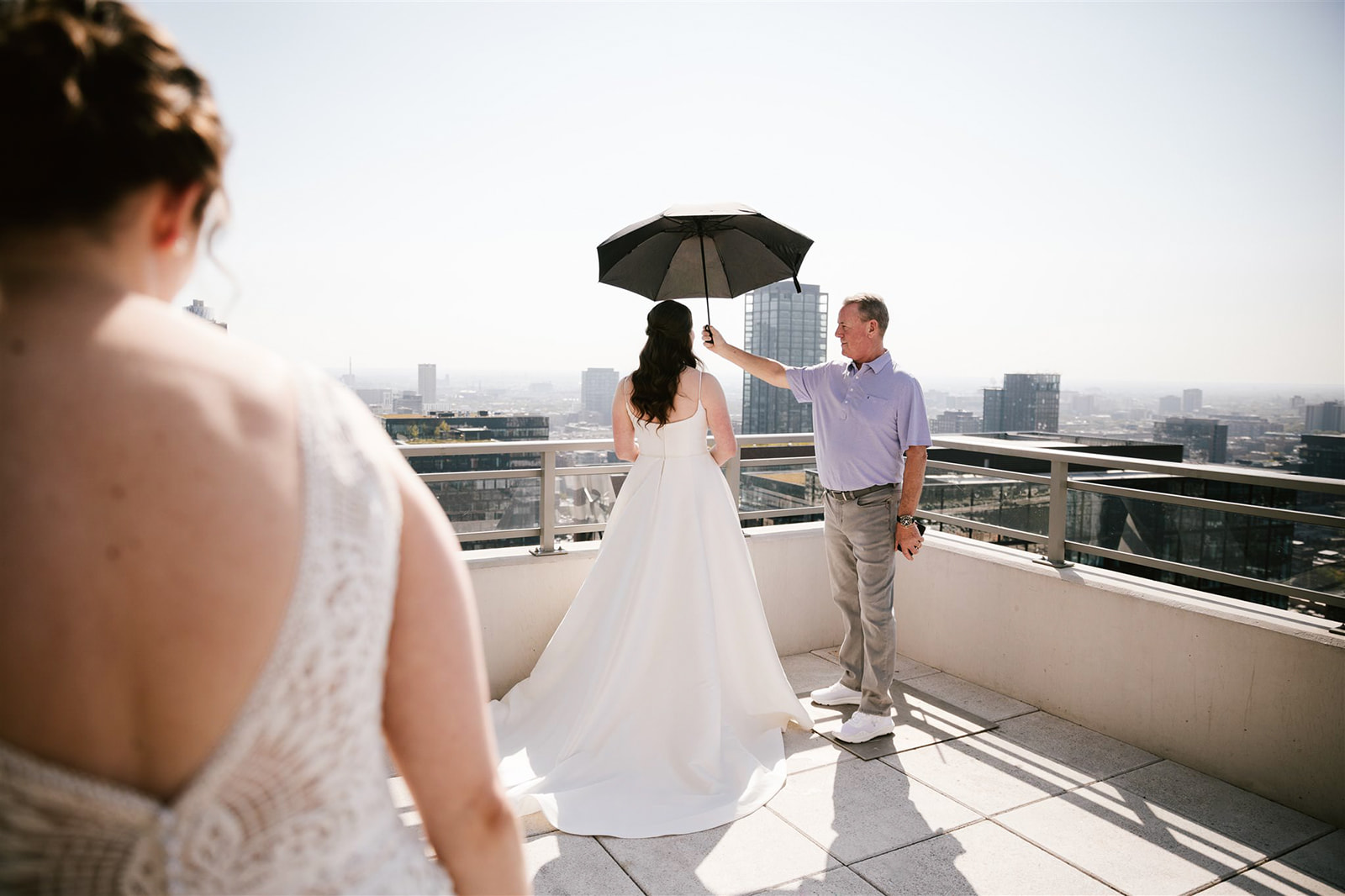 Two brides share a first look on rooftop with Chicago skyline, one bride's father holds an umbrella.