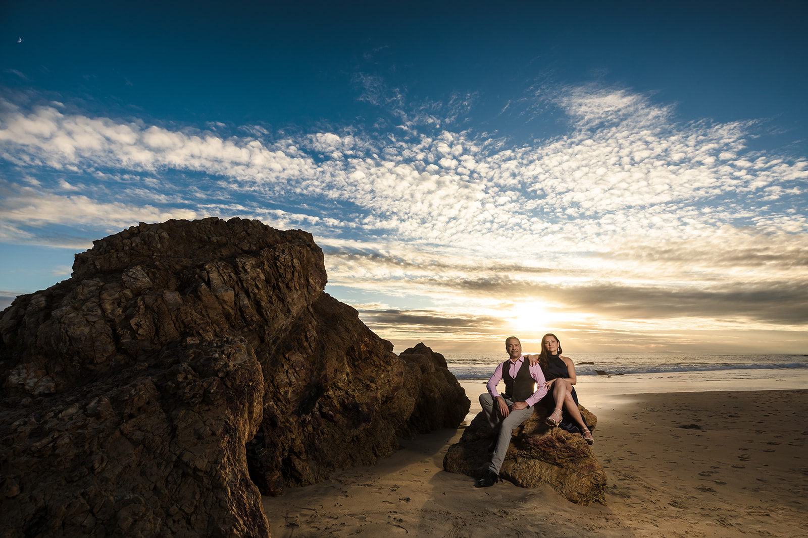 Sophisticated engaged couple sits atop a rock in front of an incredible sunset