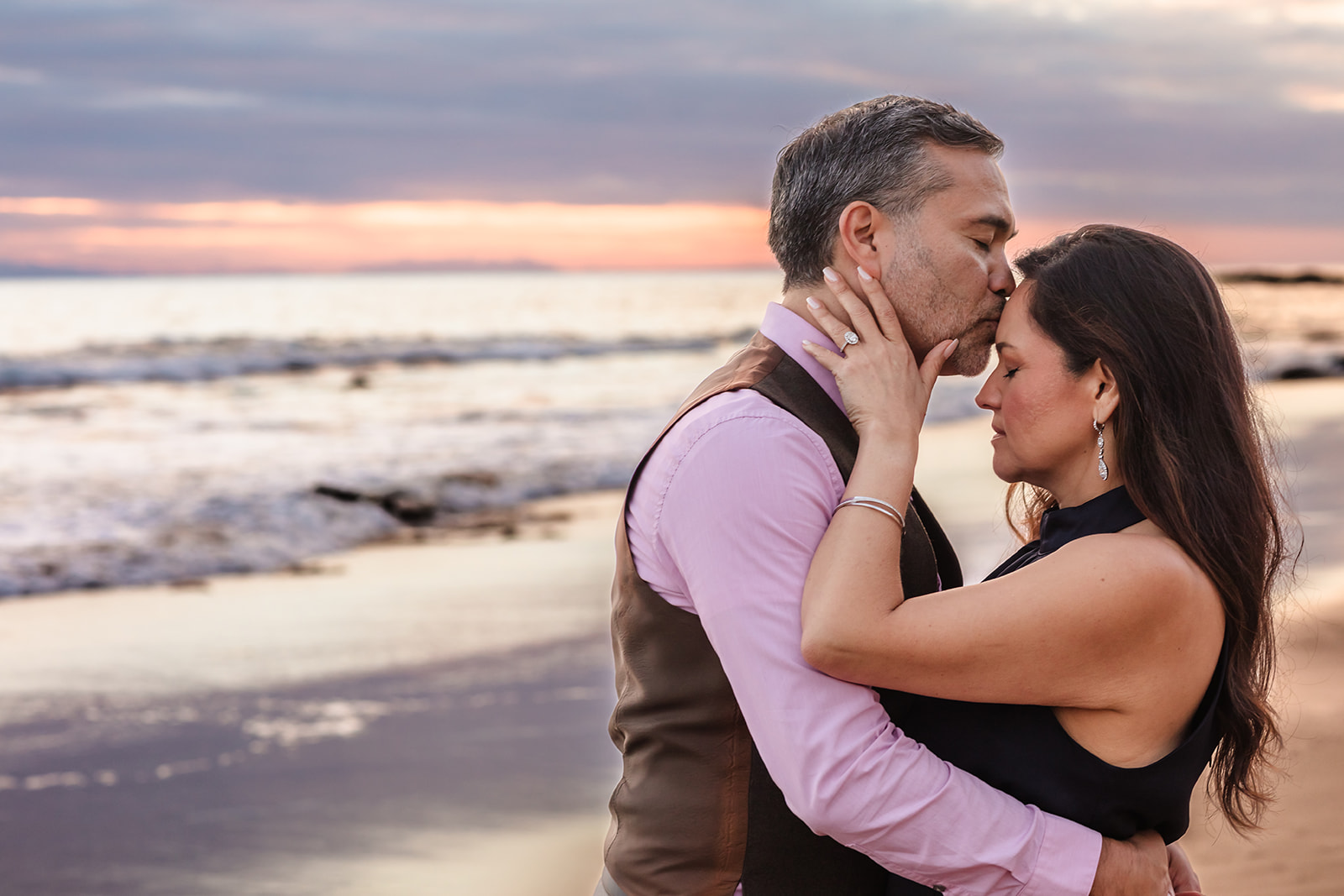 closeup enagement photo of a man kissing womans forehead in front of a pink sunset