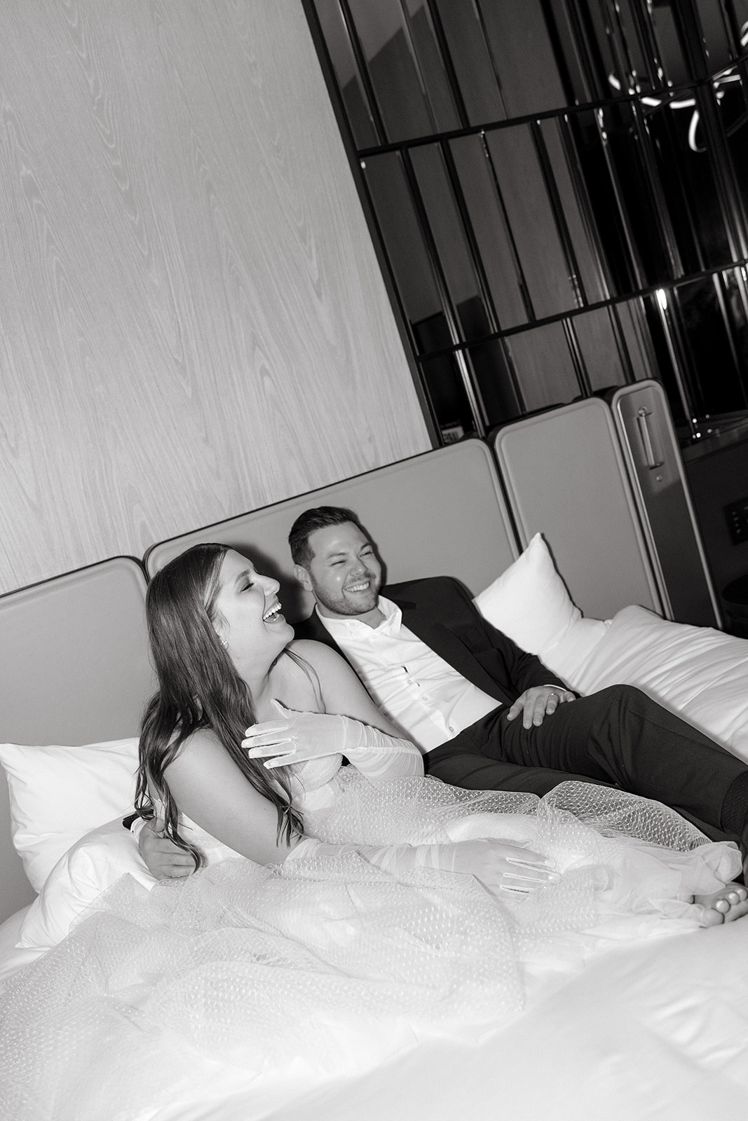 A couple who eloped in NYC sit on a hotel bed and laugh together