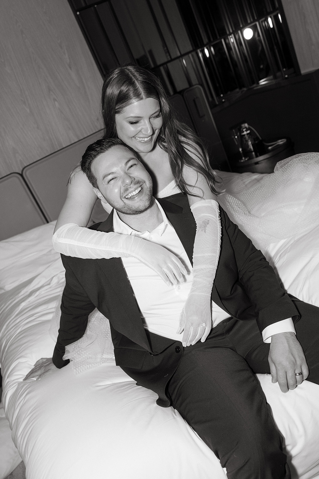 A couple who eloped in NYC sit on a hotel bed and laugh together