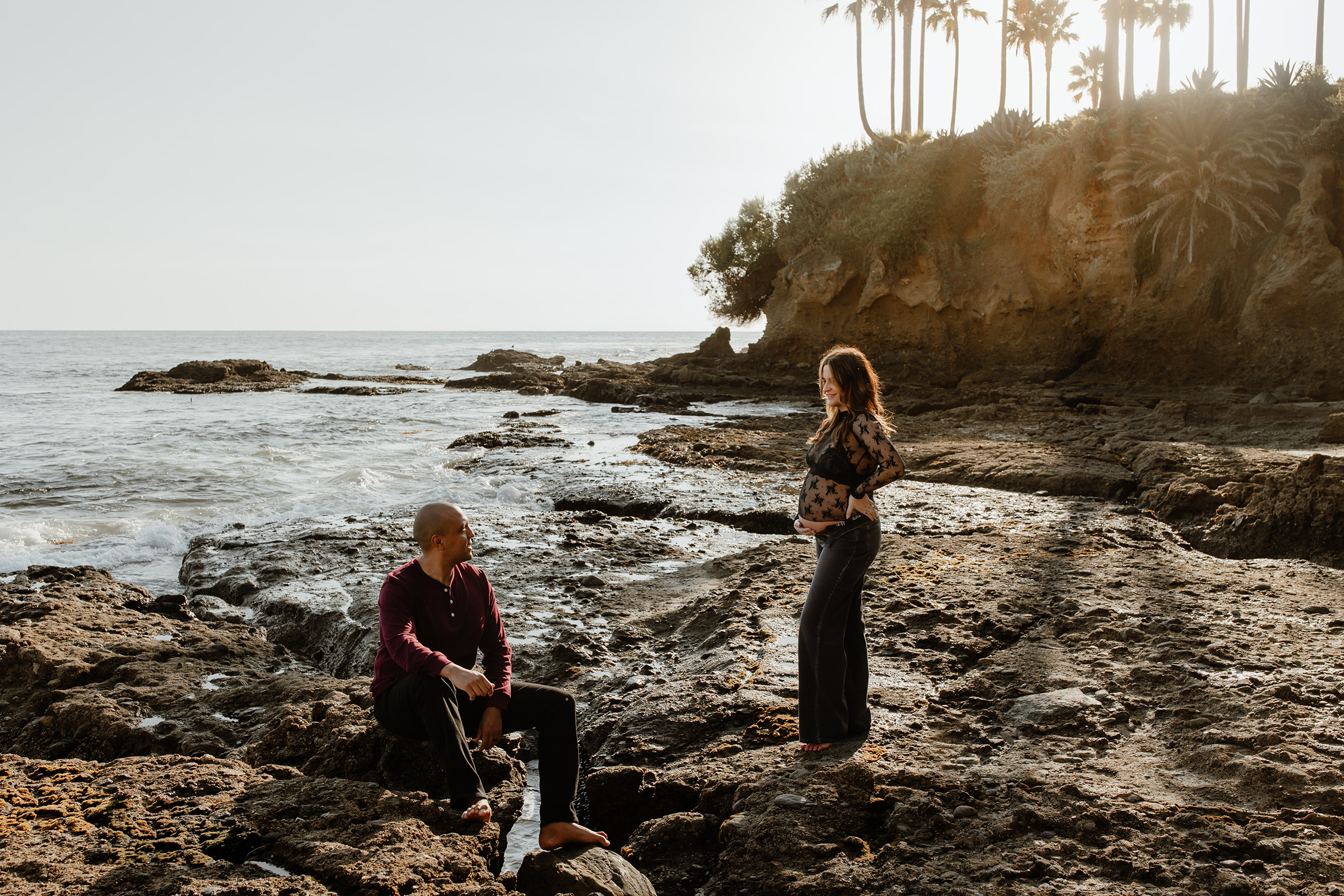 A couple at the beach for their maternity session, laguna beach, palm trees behind them, sun in the background