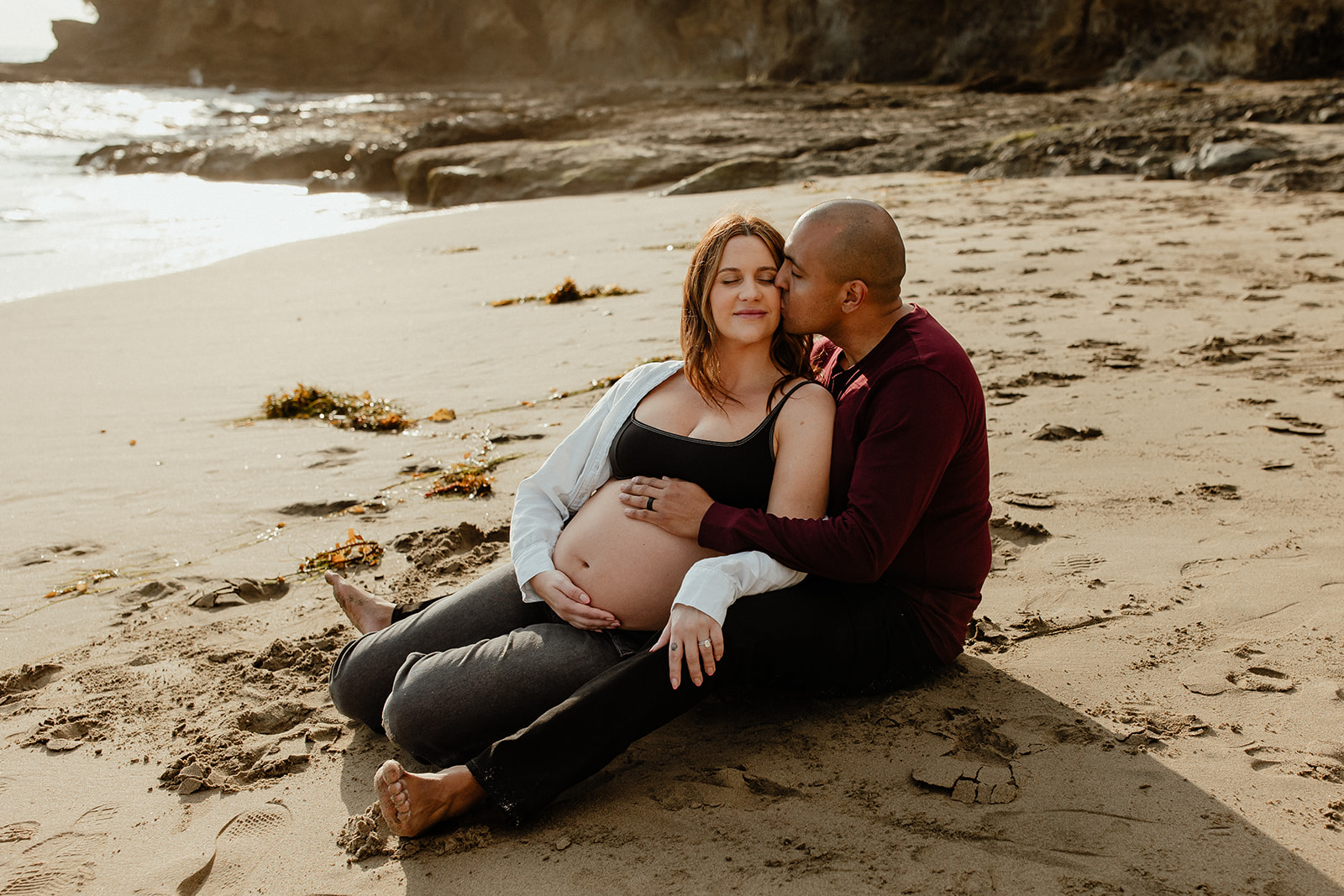 couple sitting on the sand hugging and kissing, maternity session