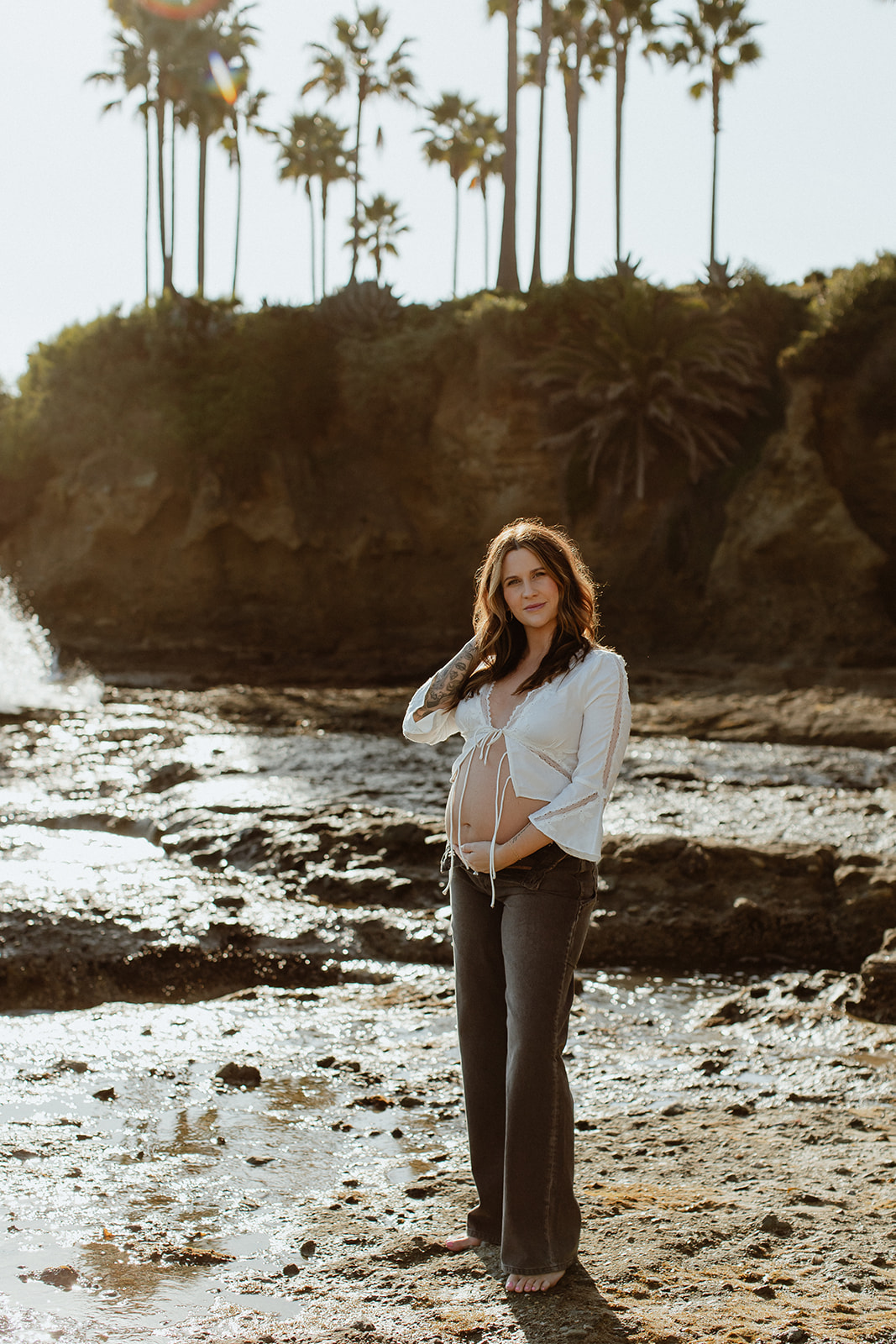 mom to be at the beach for their maternity session, laguna beach, palm trees behind them, sun in the background