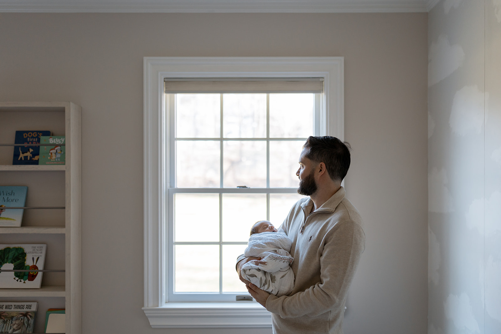 Dad holds newborn baby while looking out nursery window. 