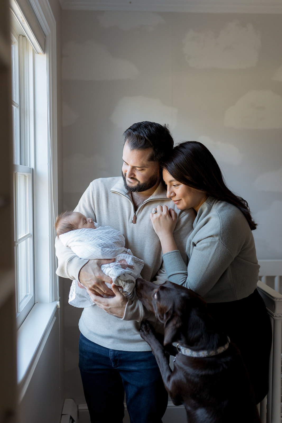 A mom and dad hold their newborn baby by the nursery window while their dog jumps up to see the baby. 
