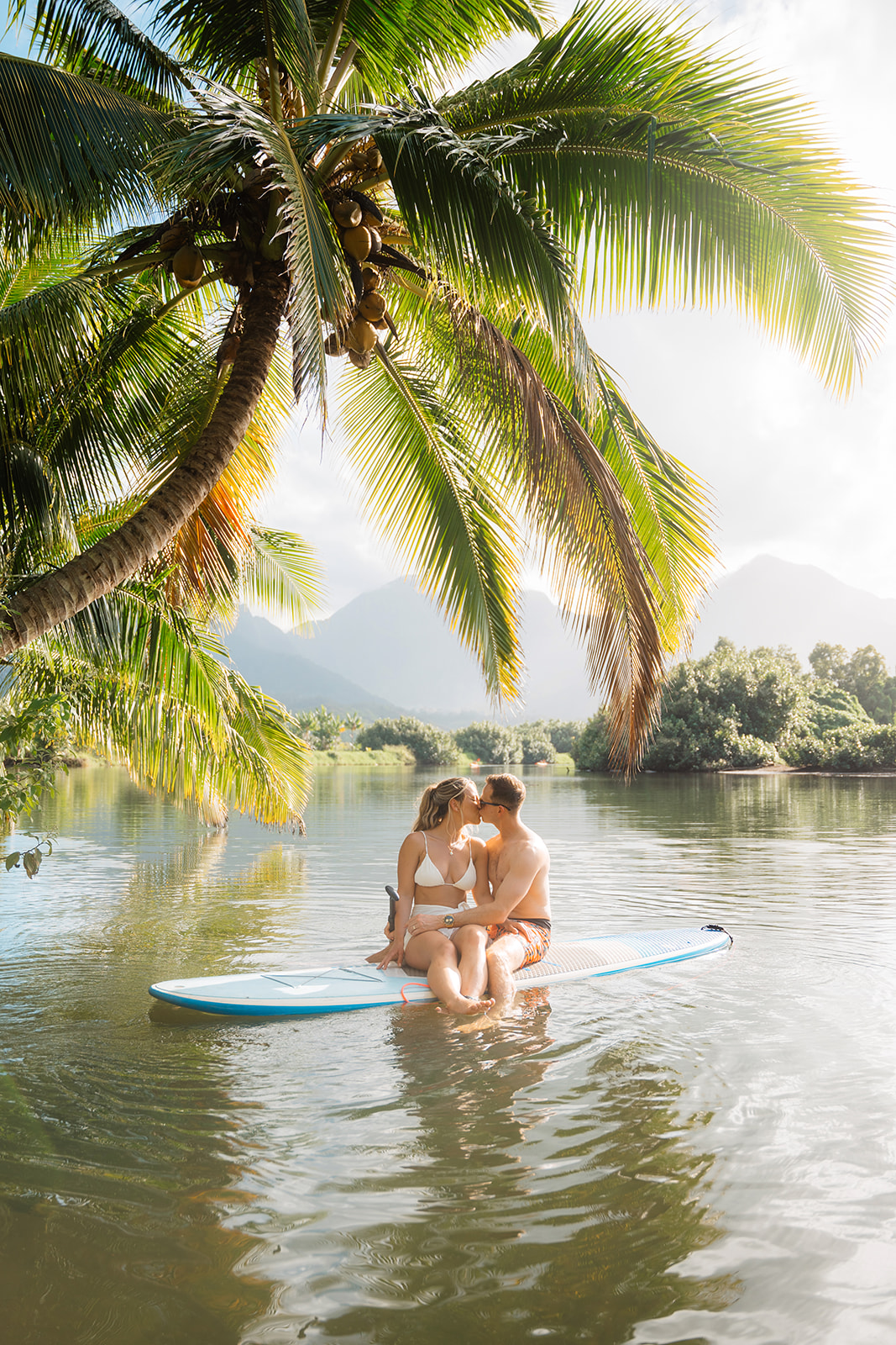 A couple kissing on a paddle board beneath a palm tree on the Hanalei river in Kauai on their elopement day. 