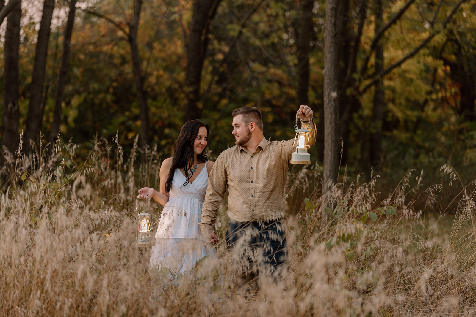 Couple holding lanterns looks at each other in the dusk while standing in tall grass