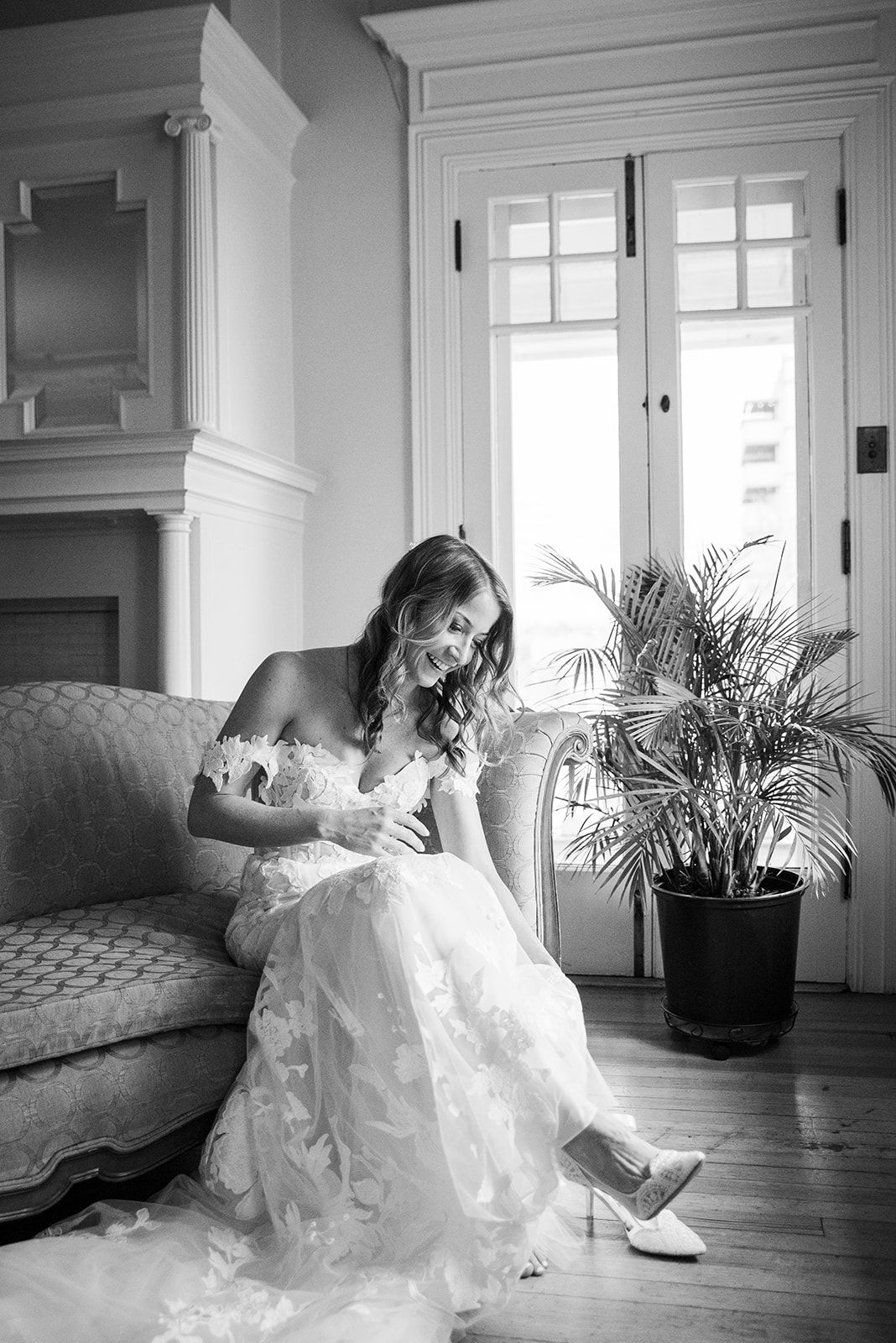 A bride sits on a vintage couch and leans over to adjust her heel, captured by Denver wedding photographer.