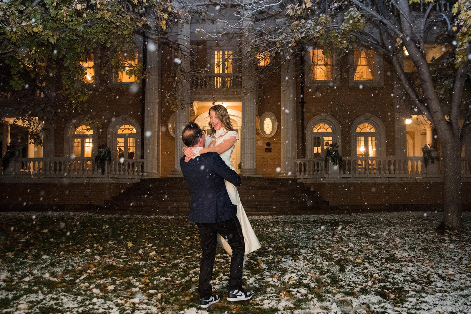 A groom lifts and spins his bride in the front lawn of the Grant Humphrey Mansion at night as snow falls around them.