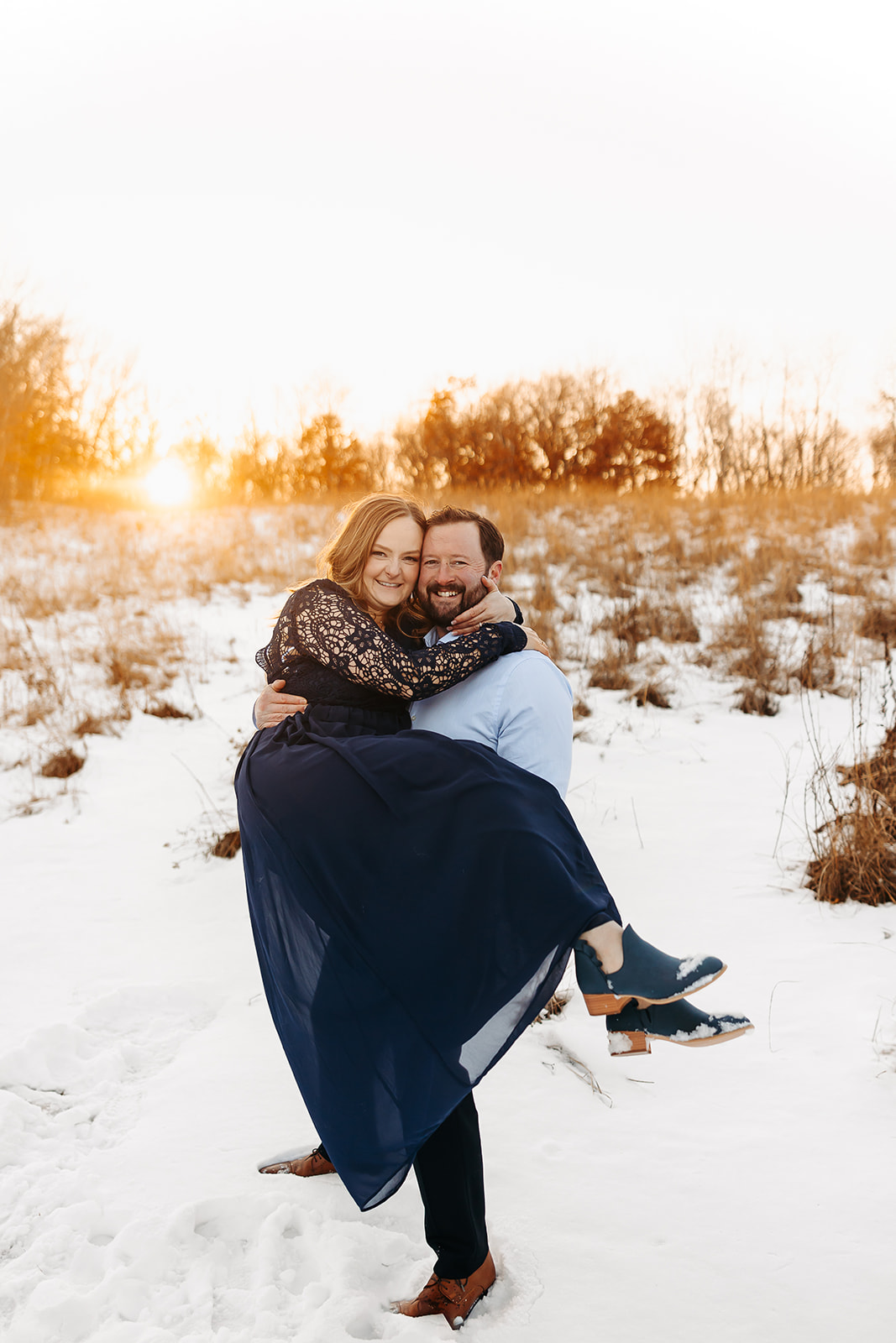 Man holding woman up in the snow while they are both smiling and the sun is behind them.