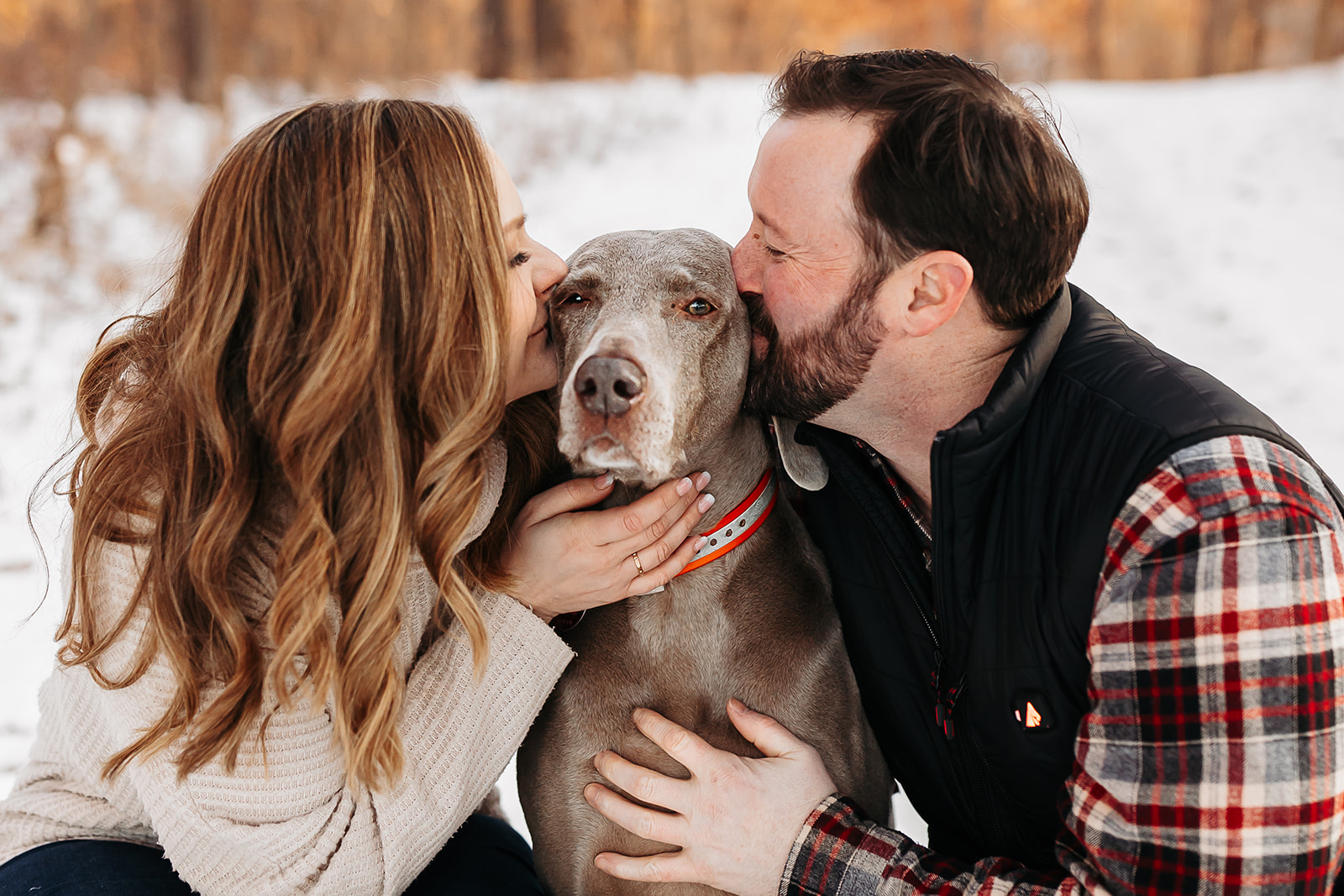 Man and woman kissing their dog which is between them in the snow.